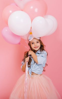 Happy celebration of birthday party with flying balloons of charming cute little girl in tulle skirt smiling to camera isolated on pink background. Charming smile, expressing happiness. Place for text